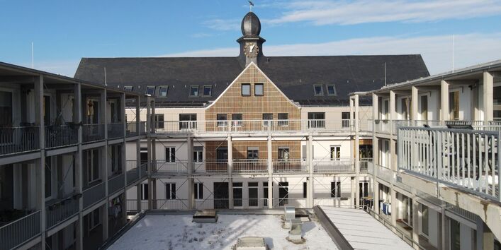 Foto eines Gebäudekomplexes mit Laubengängen. Zentralperspektive auf ein Gebäude mit schwarzem Satteldach, auf dem mittig ein kleines Türmchen mit Uhr sitzt, direkt über einem mit Holz verkleideten Zwerchgiebel. Die Gebäudeteile rechts und links wirken moderner mit Flachdächern und Betonstützen. Im Hof liegt Schnee.