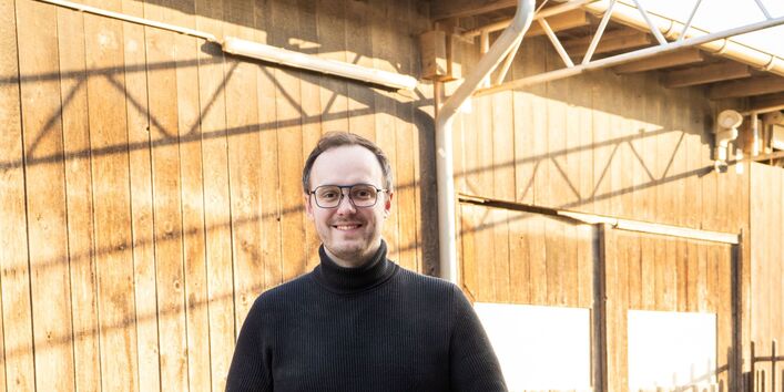 Foto eines Mannes in schwarzem Pullover hinter einer halbhohen Mauer aus Hohllochziegeln. Er hält eine Mörtelrolle und einen weiß-pinken Sack mit der Aufschrift “ClayTec Lehm-Dünnbettmörtel” in den Händen. Vor der Mauer zwei weitere Säcke.