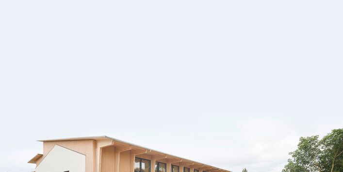 Eckansicht einer Mehrzweckhalle. Eine Holzkonstruktion kragt aus der Halle hervor und bildet einen Dachüberstand. Zwischen den Holzständern ist die Fassade vollflächig verglast. An der Giebelwand setzt sich die weiß verputzte Kontur eines ähnlich hohen Gebäudes mit Satteldach von der Holzfassade ab. Daneben eine Wiese mit einem Schotterweg und Bäumen.