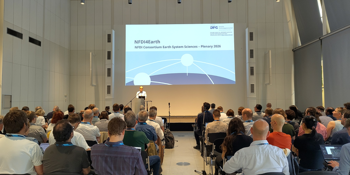 Vortragssaal mit zahlreichen Teilnehmenden bei der NFDI4Earth-Plenary 2026. Eine Person präsentiert auf der Bühne vor einer großen Leinwand mit dem Veranstaltungslogo.