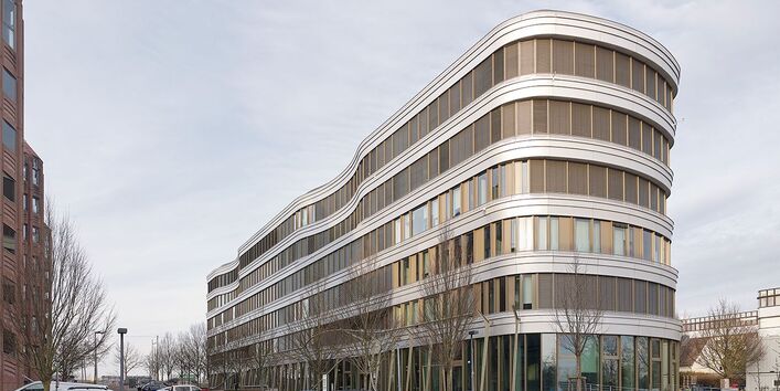 Foto eines sechsgeschossigen Bürogebäudes mit Flachdach, geschwungener Fassade, horizontal gegliederter Fensterbändern und abgerundeten Ecken. Im Vordergrund geparkte Autos, junge Bäume entlang des Gebäudes.