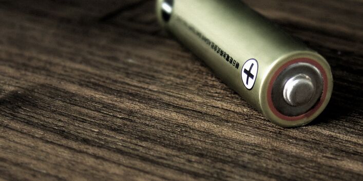 Close-up, low-angle shot of a single AA battery lying horizontally on a dark wooden surface. The focus is sharp on the positive terminal and the "plus" symbol, with the background fading into a soft blur.
