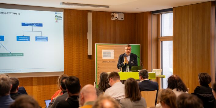 Eine Person referiert gestikulierend in einem Sitzungsraum. Im Vordergrund sind unscharf die Hinterköpfe des Publikums abgebildet. Im Hintergrund wird eine Präsentation projiziert.