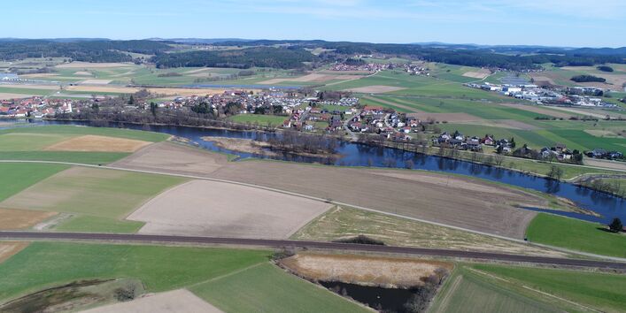 Luftbildaufnahme des Flusses Naab mit Grünland- und Ackerflächen, die bis an die Gewässerkante reichen. Parallel zum Fluss verlaufen auf beiden Seiten Verkehrswege.