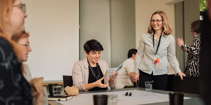 Eine Gruppe Workshop Teilnehmerinnen sitzen um einen Tisch. Darauf liegt großformatiges Papier, auf dem Workshop Ergebnisse notiert werden.