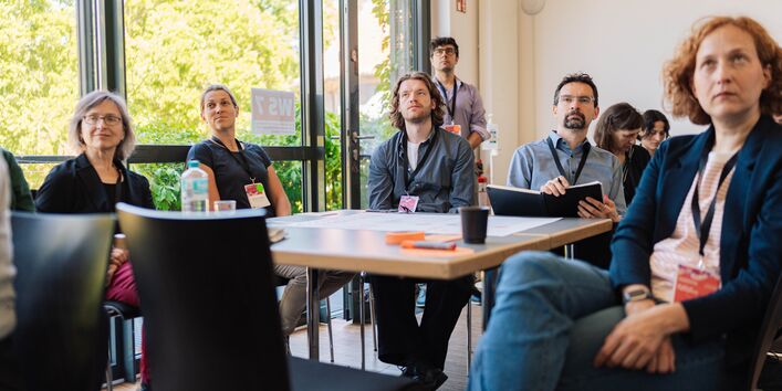 Die Teilnehmenden sitzen an einem Tisch und lauschen einem Vortrag.