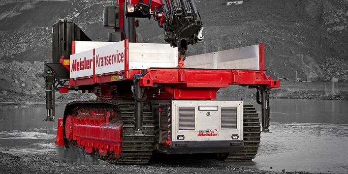 auf einem selbstfahrenden Raupenfahrzeug wird ein Kran durch bergiges Gelände und flaches Wasser transportiert