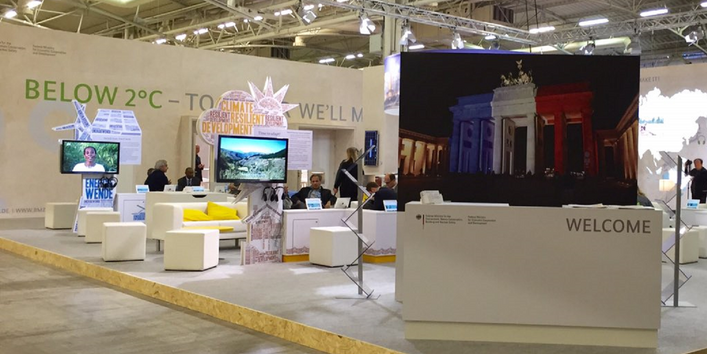 in einer Messehalle sind Stellwände, Sitzgelegenheiten und Bildschirme aufgebaut. Eine Stellwand zeigt das Brandenburger Tor, das in den Farben der französischen Flagge angestrahlt ist. Auf einer Stellwand hinten steht "Below 2 °C - together we´ll make it