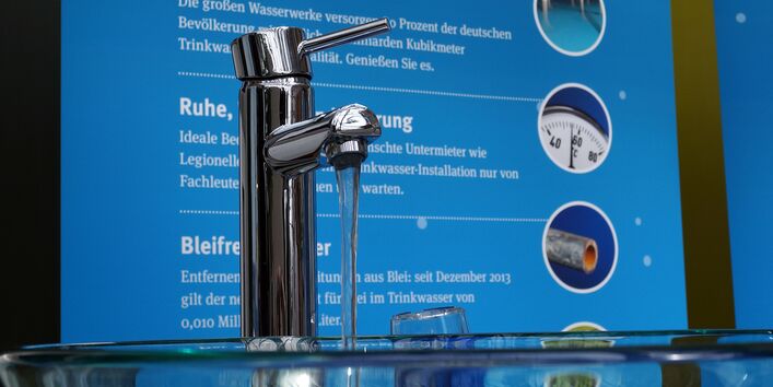 an einem Informationsstand hängt ein Plakat mit Verbrauchertipps rund um das Trinkwasser, aus einem Wasserhahn läuft Wasser in eine Glasschüssel
