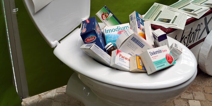 An einem Infostand steht ein Toilettenbecken, in dem sich Medikamentenschachteln stapeln. Auf dem aufgeklappten Klodeckel klebt ein Zettel: eine Hand, die Tabletten ins Klo wirft, ist rot durchgestrichen.
