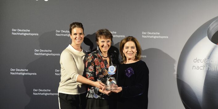 drei Frauen stehen lächelnd auf einer Bühne und halten eine "Blauer Engel"-Trophäe in die Kamera, im Hintergrund eine Wand mit Schriftzügen "Der Deutsche Nachhaltigkeitspreis"