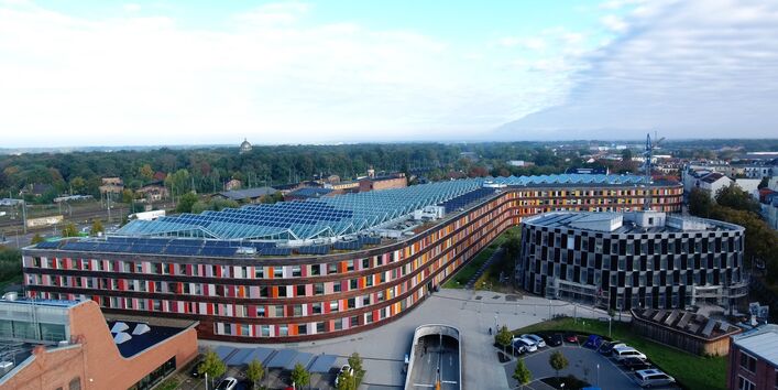 Foto von schräg oben vom Hauptgebäude des UBA Dessau mit seiner Fassade aus Holz und buntem Glas und einer Photovoltaikanlage auf dem Dach und daneben der Erweiterungbau mit seiner Fassade aus Photovoltaikmodulen