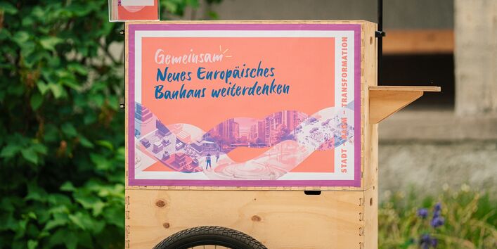 Holzstand mit Plakat zur Konferenz 'Gemeinsam. Neues Europäisches Bauhaus weiterdenken' mit den Worten 'Stadt – Raum – Transformation' und städtischen Illustrationen