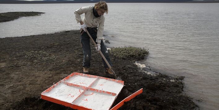 Die Künstlerin steht an Ufer eines Sees und nimmt eine Bodenprobe. 