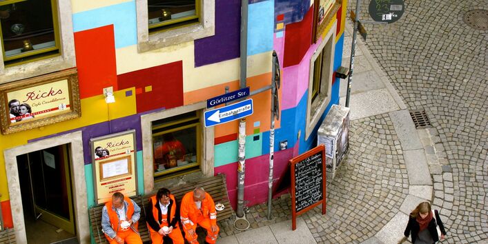 Drei Männer in den orangenen Anzügen der Müllabfuhr sitzen auf einer Bank vor einem bunt gestrichenen Eckhaus und trinken Kaffee
