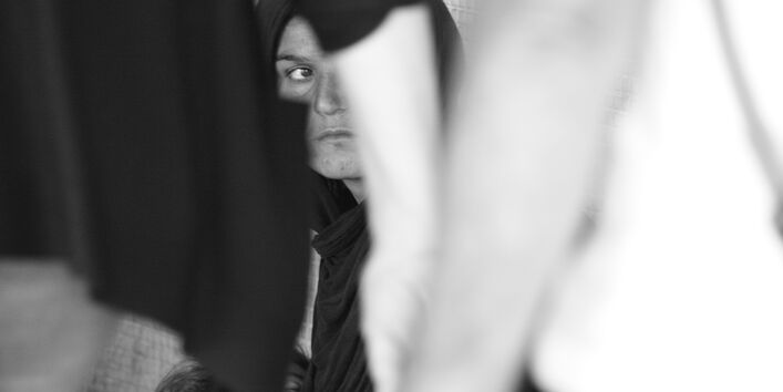 Eine Frau sitzt mit ihrem Kind im Arm am Straßenrand, zwei Passanten gehen an ihnen vorbei.