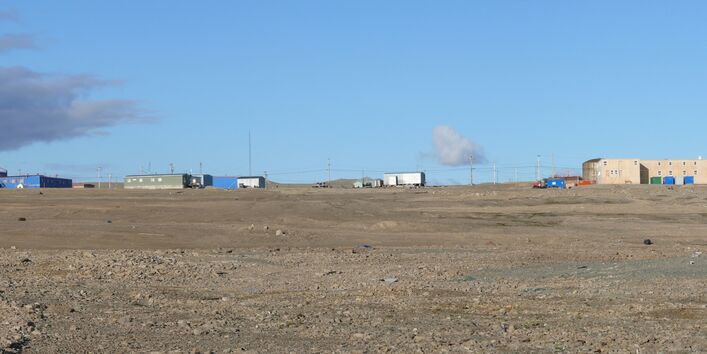 Im Hintergrund stehen einige Hallen und Häuser, vorne ist die karge Landschaft der Arktis zu sehen. 