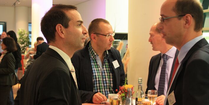 Vier Männer stehen an einem Stehtisch. Jeweils zwei unterhalten sich angeregt miteinander. Zwei tragen eine Brille und alle tragen Anzug und Hemd. Auf dem Tisch steht eine kleine Vase mit einer Blume.