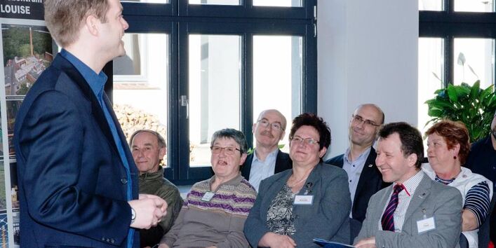 Ein Mann im Anzug steht vor einigen Personen im Publikum und spricht. Er hat die Hänge zusammen gefaltet und schaut geradeaus. Im Publikum sitzen 7 Personen in zwei Stuhlreihen hintereinander und hören zu. Sie tragen Namensschilder