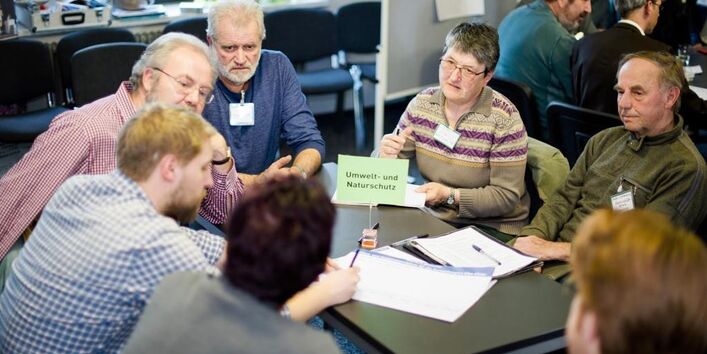 Sieben Personen sitzen um einen schwarzen Tisch. Sie tragen Namensschilder und diskutieren angeregt. Auf dem Tisch liegen verschieden Zettel. Eine Person füllt ein Blatt aus.