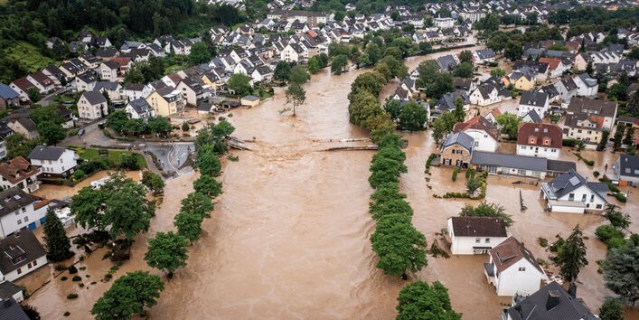 The picture shows a housing estate with single- and small multi-family houses along a river valley. The river flowing through the village has burst its banks and its brownish water has inundated the low-lying parts of the village. The original outline of the riverbed is indicated by rows of trees on what normally is the river bank. In the centre of the picture, you can see a destroyed bridge which used to cross the river. 