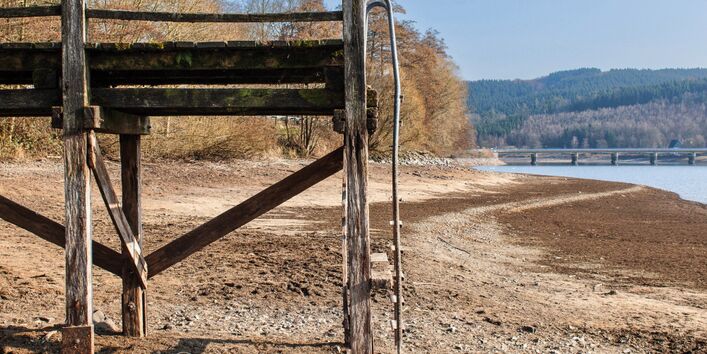 The picture shows a footbridge leading across the dried-up sandy/stony shore area of a lake. A bridge and some residual water are visible in the background.