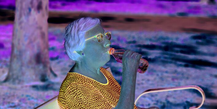 The picture shows an elderly woman sitting on a park bench in the shade of a tree, taking a drink from a bottle of water. 