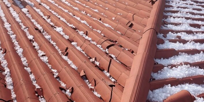 The picture shows the top view of a building roofed with red tiles. To the right and left of the roof ridge you can see numerous large hailstones. Some of the roof tiles are broken. 