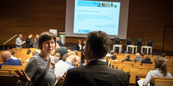 Blick von hinten in den Hörsaal des Umweltbundesamtes. Auf den Plätzen sitzen Veranstaltungsteilnehmerinnen und -teilnehmer und schauen auf eine Präsentation nach vorne. Zwei Personen diskutieren im Vordergrund.
