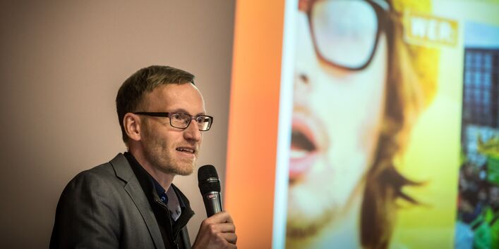 Vortragender steht mit Mikrofon in der Hand neben seiner Präsentation.