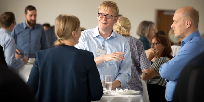 Teilnehmerinnen und Teilnehmer stehen während der Pause unterhaltend beisammen. Ein Teilnehmer hält ein Wasserglas in der Hand und spricht.