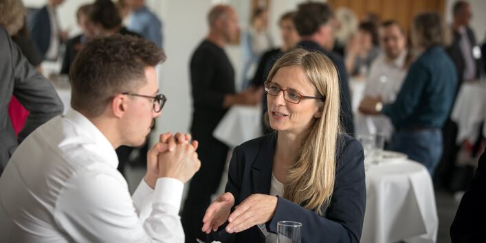 Teilnehmer und Teilnehmer stehen gemeinsam am Bistrotisch und unterhalten sich gestikulierend miteinander. Im Hintergrund sind stehen andere Teilnehmerinnen und Teilnehmer ebenfalls um den Bistrotischen herum.
