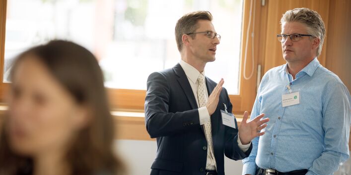 Zwei Teilnehmer stehen unterhaltend vor einem großen Fenster während der Pause am Bistrotisch.