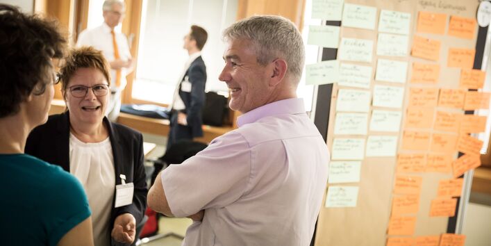 Drei Teilnehmerinnen und Teilnehmer stehen zusammen und schauen sich freundlich an und sprechen miteinander. Seitlich befindet sich eine mit Moderationskarten bestückte Moderationswand. Im Hintergrund sieht man ebenfalls Teilnehmer vor einem Fenster stehen, die sich miteinander sprechen.