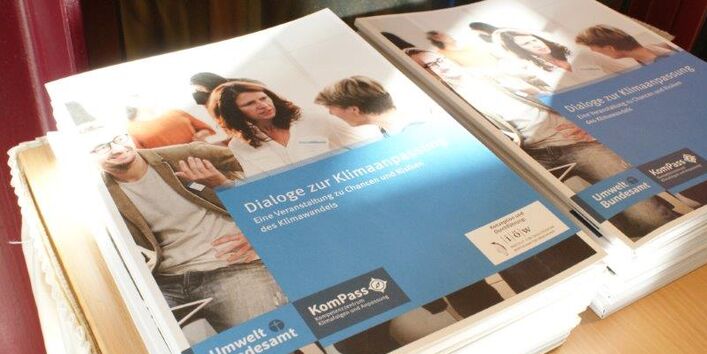 Draufsicht auf zwei Stapel der Broschüre mit dem Titel: Dialoge zur Klimaanpassung - Eine Veranstaltung zu Chancen und Risiken des Klimawandels. Auf dem Cover diskutieren gestikulierend Personen miteinander. Die Logos der Veranstalter sind ebenfalls abgebildet.