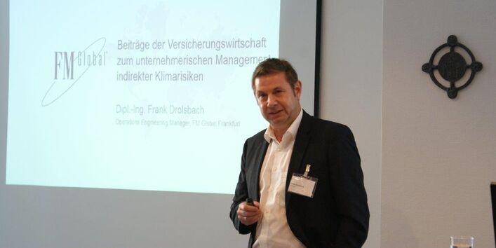 Vortragender steht mit dem Rücken vor seiner Präsentation im Hintergrund. Er hält einen Pointer in seiner Hand und spricht zu den Teilnehmern.