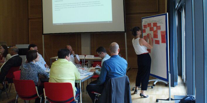 Teilnehmende präsentiert an Moderationswand mit roten Moderationskarten. Die anderen Teilnehmenden sitzen nachdenklich am Gruppenarbeitstisch. Im Hintergrund ist eine Präsentation eingblendet.