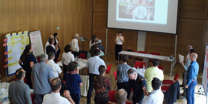 Alle Teilnehmenden stehen im Raum und folgen der Präsentation einer Teilnehmerin, die vor der Leinwand steht. Manche Teilnehmende applaudieren. Teile der Präsentationswände mit den farbigen Moderationskarten stehen am Rand verteilt