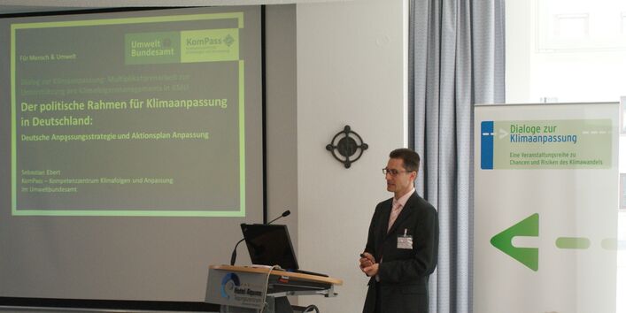 Vortragender steht am Rednerpult. Im Hintergrund sieht man die Präsentation auf Projektionswand und Veranstaltungsaufsteller.