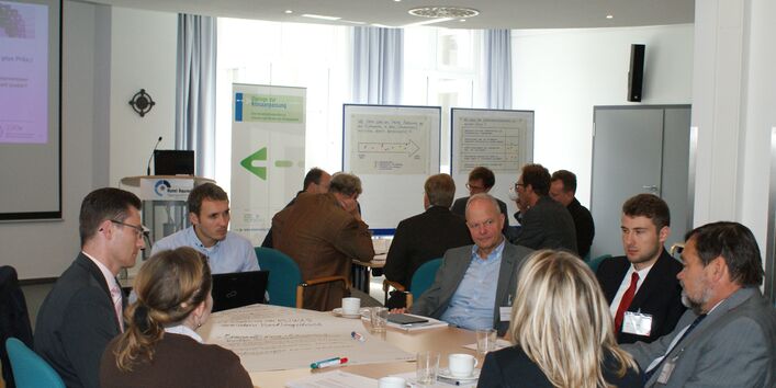 Teilnehmer sitzen an Gruppenarbeitstischen und sprechen miteinander. Im Hintergrund stehen Moderationswände vor der Fensterfront.