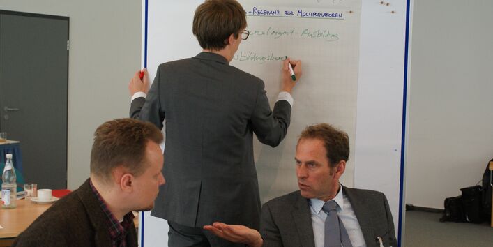 Teilnehmer schreibt auf Papier an Moderationswand. Im Vordergrund sitzen zwei Teilnehmer im Gespräch.