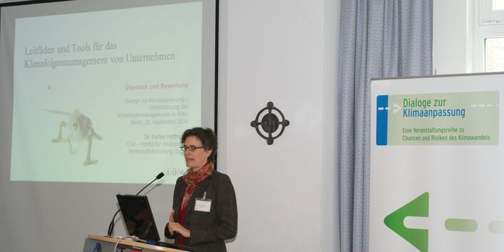 Vortragende steht am Rednerpult und trägt vor. Im Hintergrund sieht man den Vortrag auf der Projektionswand und den Veranstaltungsaufsteller.