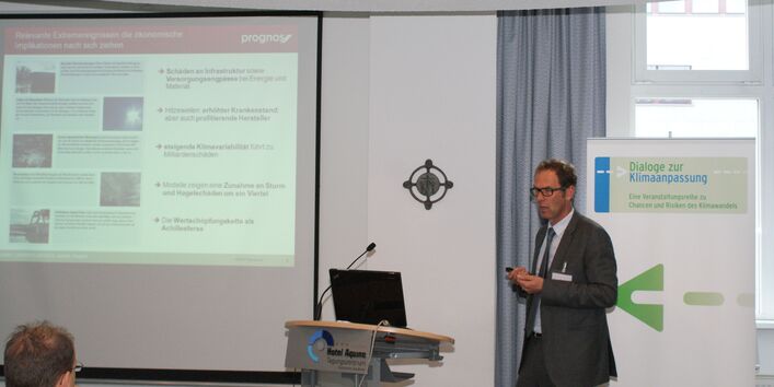 Vortragender steht am Stehpult. Im Hintergrund ist der Vortrag auf Projektionswand und Veranstaltungsaufsteller zu sehen.