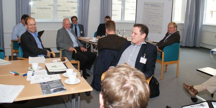 Teilnehmer sitzen an Gruppenarbeitstischen und lachen. Im Hintergrund steht eine Moderationswand vor der Fensterfront.
