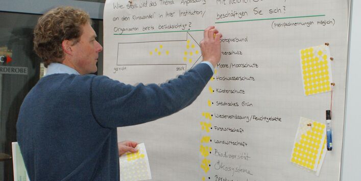 Teilnehmer steht vor Moderationswand und klebt gelbe Bewertungspunkte an zum Thema: Wie stark die Anpassung an den Klimawandel in der Institution verankert ist und mit welchen Themen der Teilnehmer befasst ist.