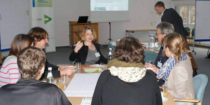 Teilnehmer sitzen am Gruppenarbeitstisch und hören einer Teilnehmerin bei ihrem Redebeitrag zu. Im Hintergrund sieht man den Veranstaltungsaufsteller, das Rednerpult und die Projektionswand und einen anderen Teilnehmer stehen.