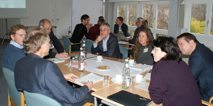 Teilnehmer sitzen an Gruppenarbeitstischen im Raum vor einer hellen Fensterfront und stehen im Dialog miteinander.