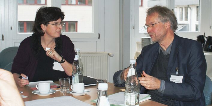 Zwei Teilnehmer sitzen am Gruppenarbeitstisch im Dialog miteinander vor einer hellen Fensterfront.