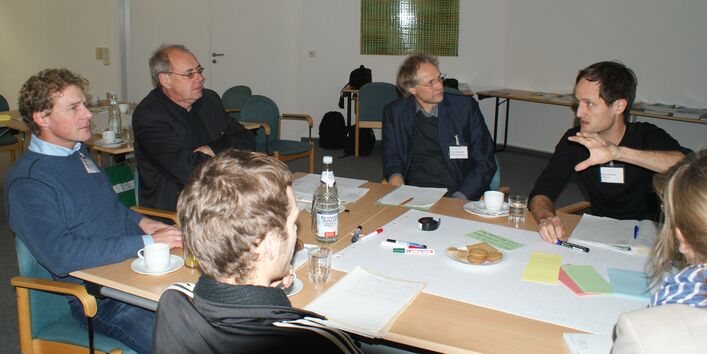 Teilnehmer sitzen am Gruppenarbeitstisch und hören einem anderen Teilnehmer zu.