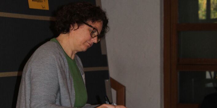 Teilnehmende steht am Stehtisch und beschreibt eine gelbe Moderationskarte. Im Hintergrund steht eine Moderationswand. Auf dieser Moderationswand sind Moderationskarten in gelb, grün, orange aufgepinnt.